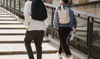 Two people with backpacks walking up outdoor concrete steps beside a metal railing, carrying laptops in a city setting.