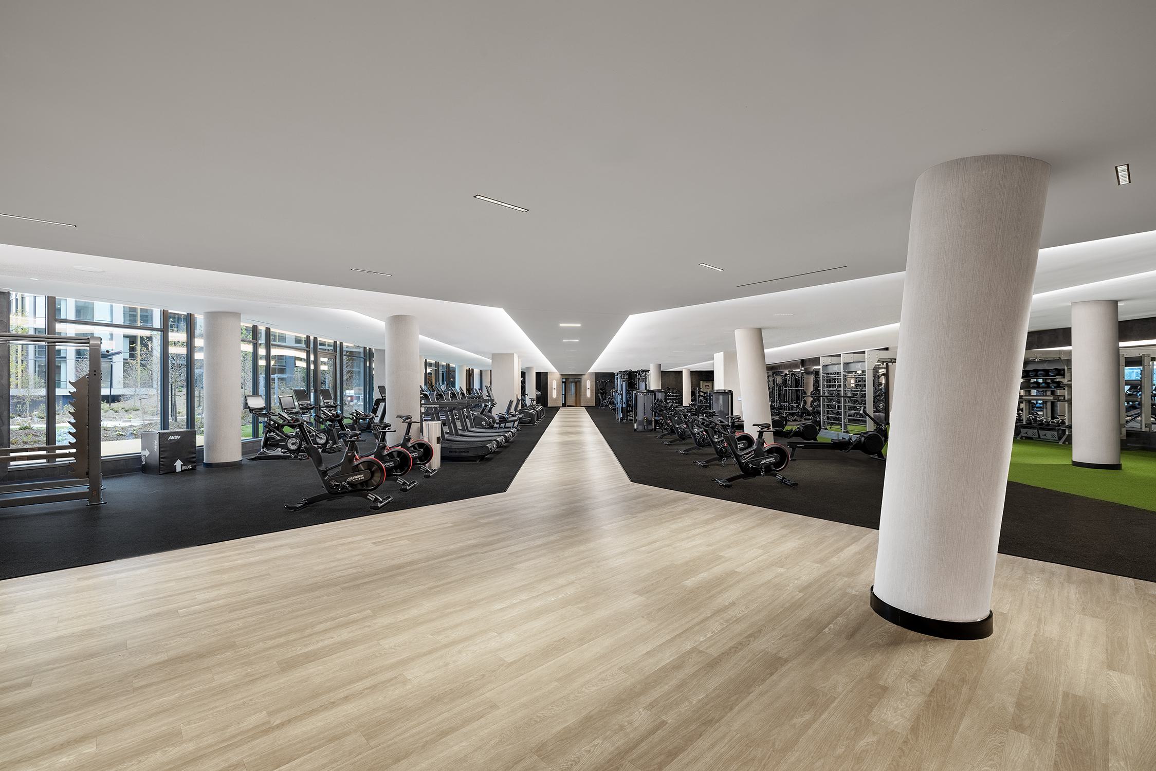 Wide-angle view of a modern fitness center featuring cardio machines on one side, strength equipment on the other, a central wood-look corridor, and high ceilings with linear lighting.
