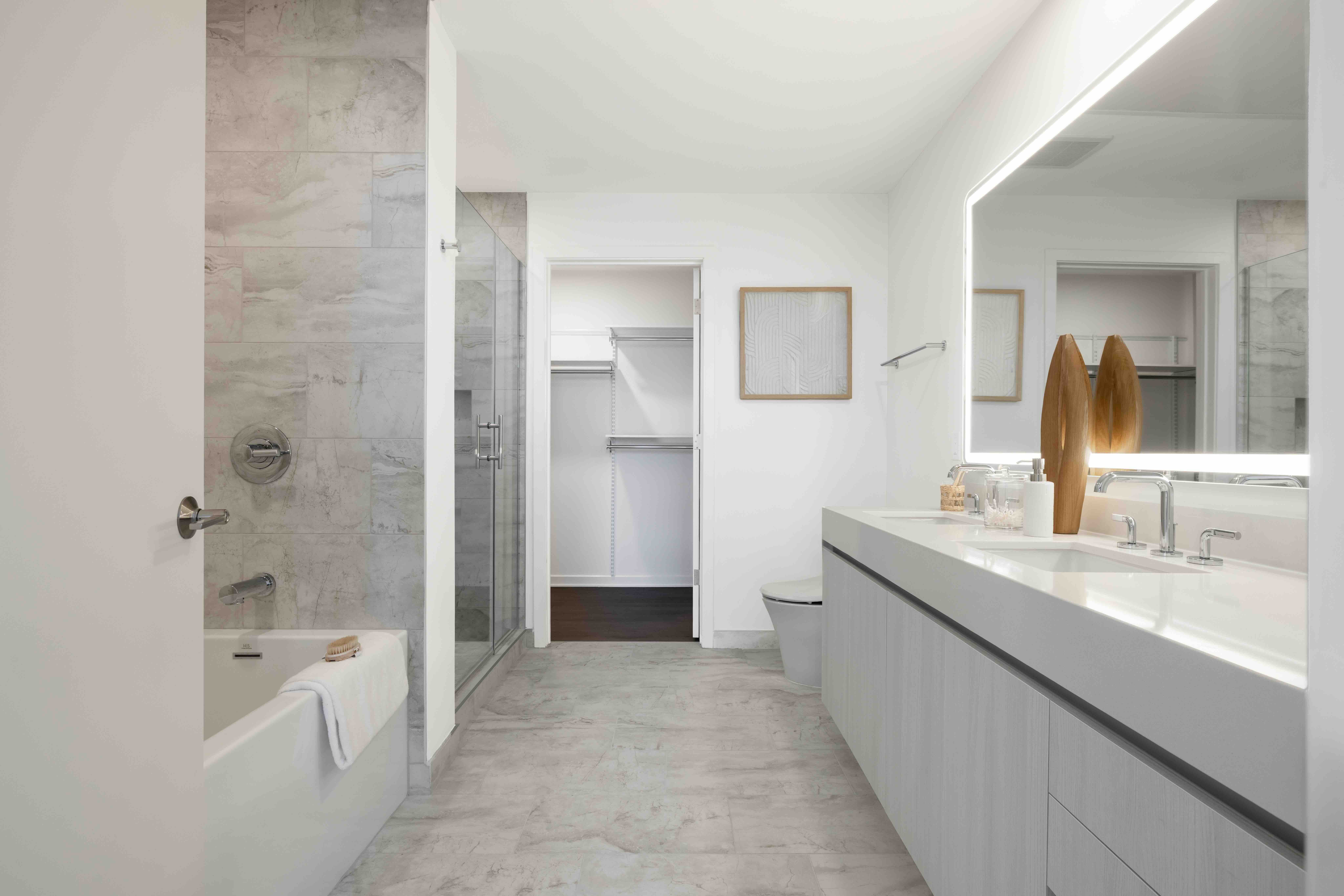 A modern penthouse bathroom featuring a long floating vanity, large backlit mirror, glass shower, and alcove bathtub.