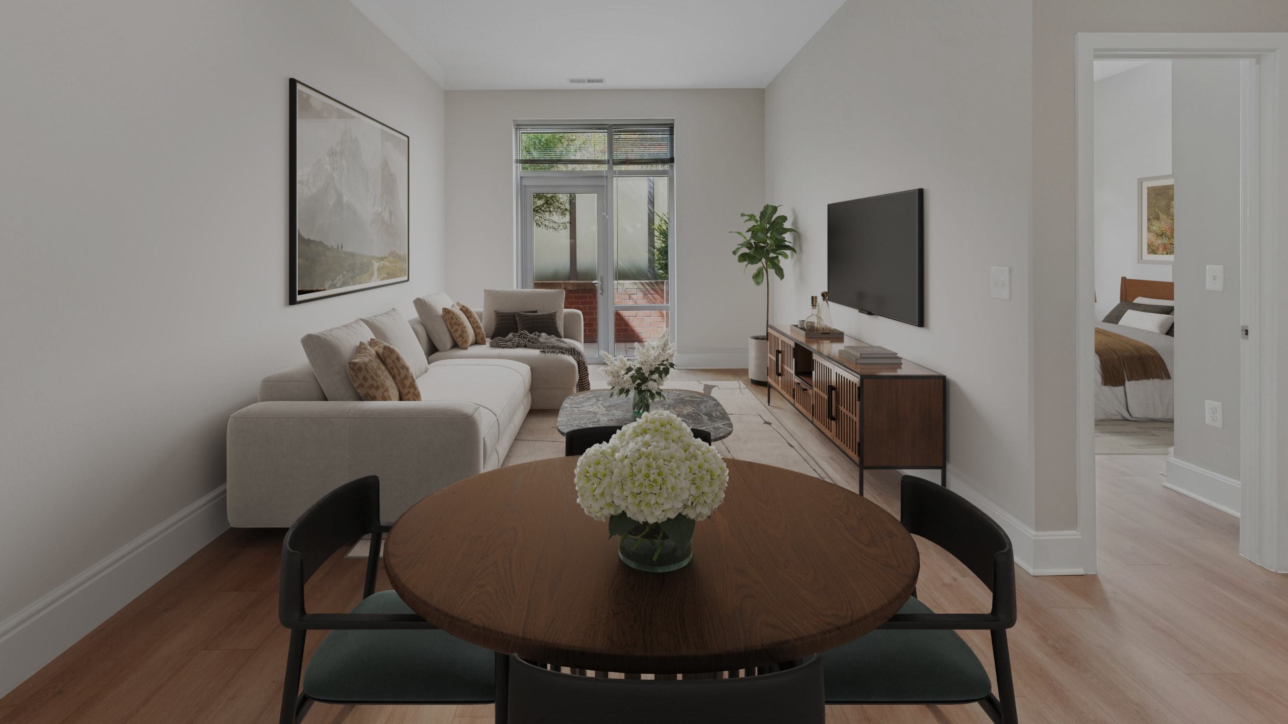 Contemporary open-plan apartment view from a dining table, showing a living area with a light grey sectional couch, a wall-mounted TV, a wooden console media cabinet, and a glass patio door leading outside.