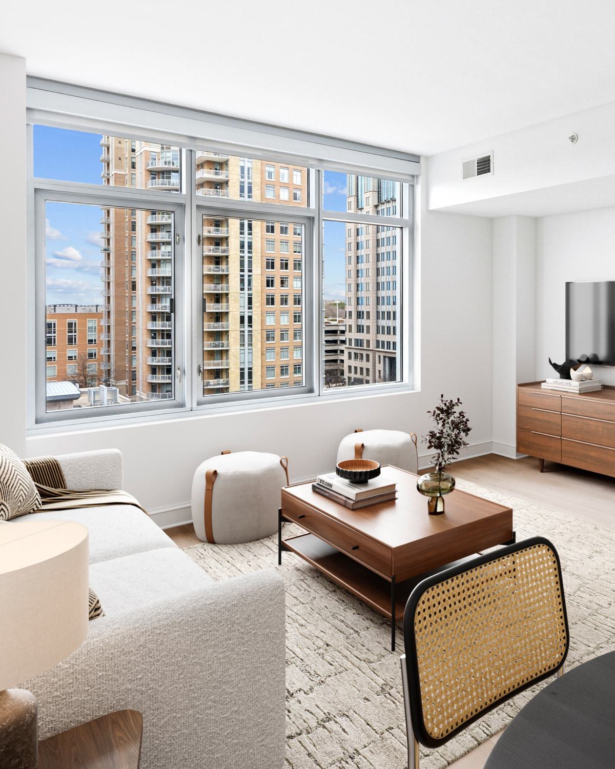 Modern, open-concept apartment living room and kitchen with light wood cabinets, a grey sofa, large windows showing city high-rises, and a wall-mounted TV.
