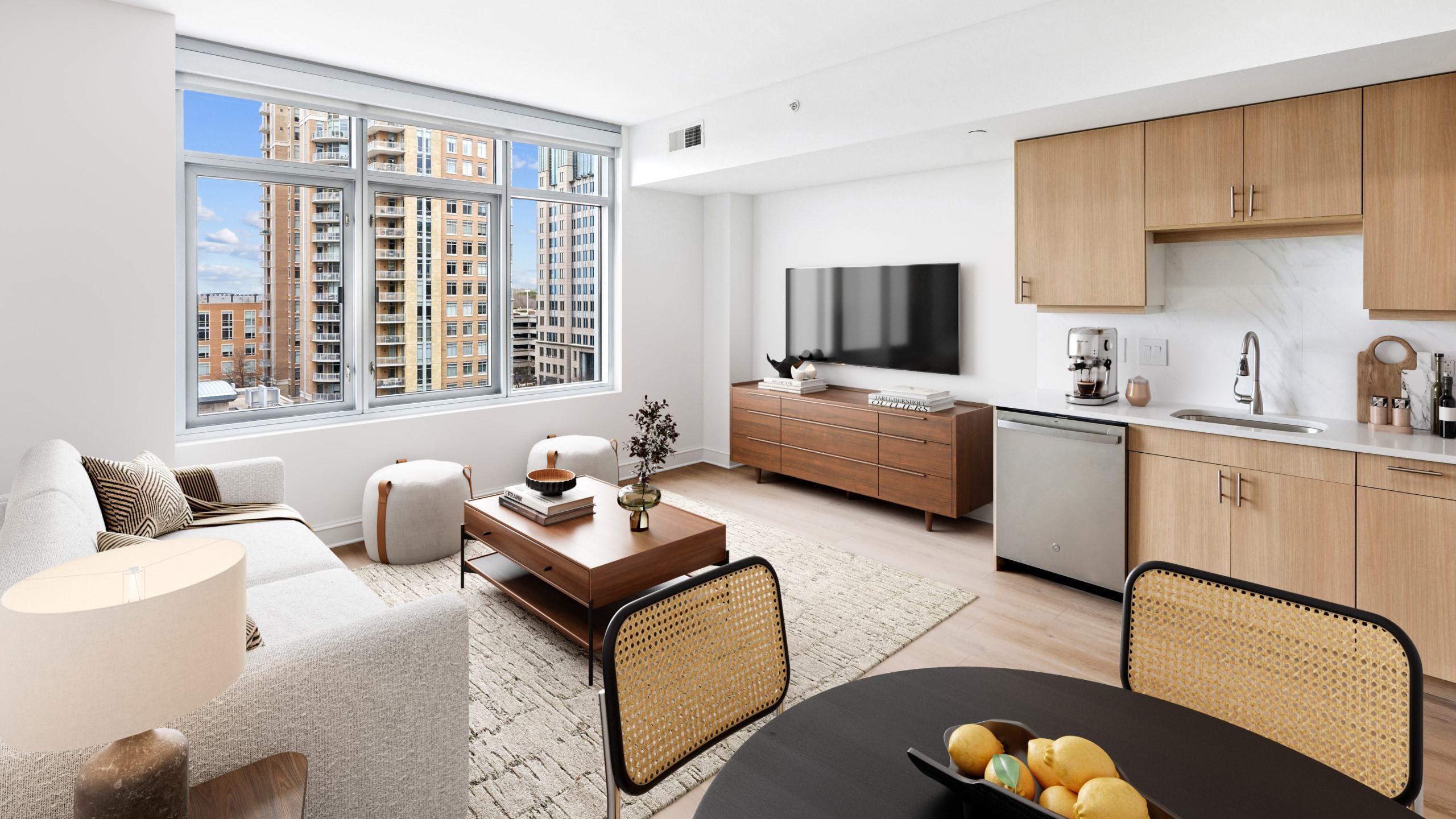 Modern, open-concept apartment living room and kitchen with light wood cabinets, a grey sofa, large windows showing city high-rises, and a wall-mounted TV.