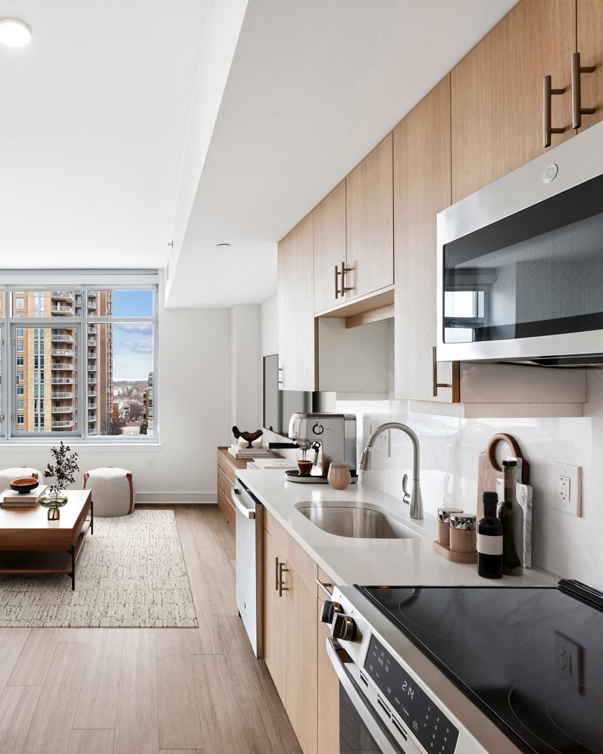 

Modern, open-concept apartment living room and kitchen with light wood cabinets, a grey sofa, large windows showing Reston Town Center high-rises, and a wall-mounted TV.