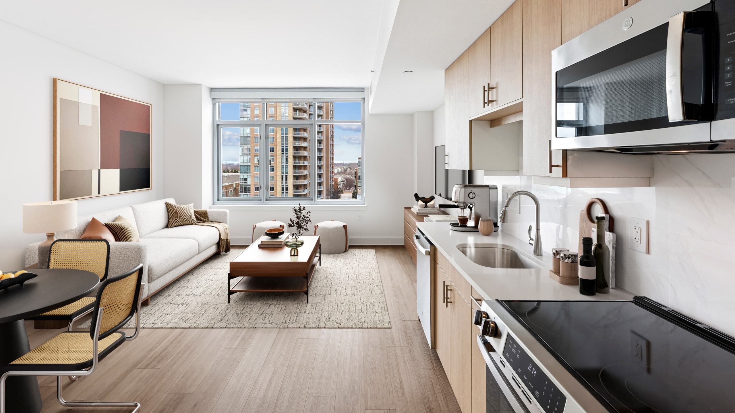 

Modern, open-concept apartment living room and kitchen with light wood cabinets, a grey sofa, large windows showing Reston Town Center high-rises, and a wall-mounted TV.