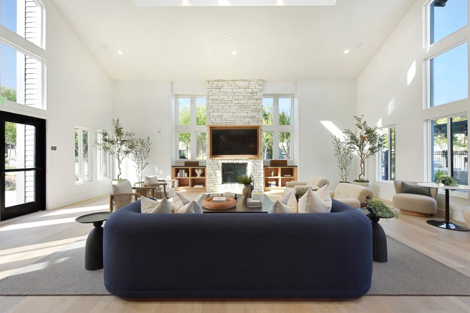 A spacious, sunlit clubhouse with high vaulted ceilings, a prominent stone fireplace, and modern furnishings anchored by a large, curved navy blue sofa.