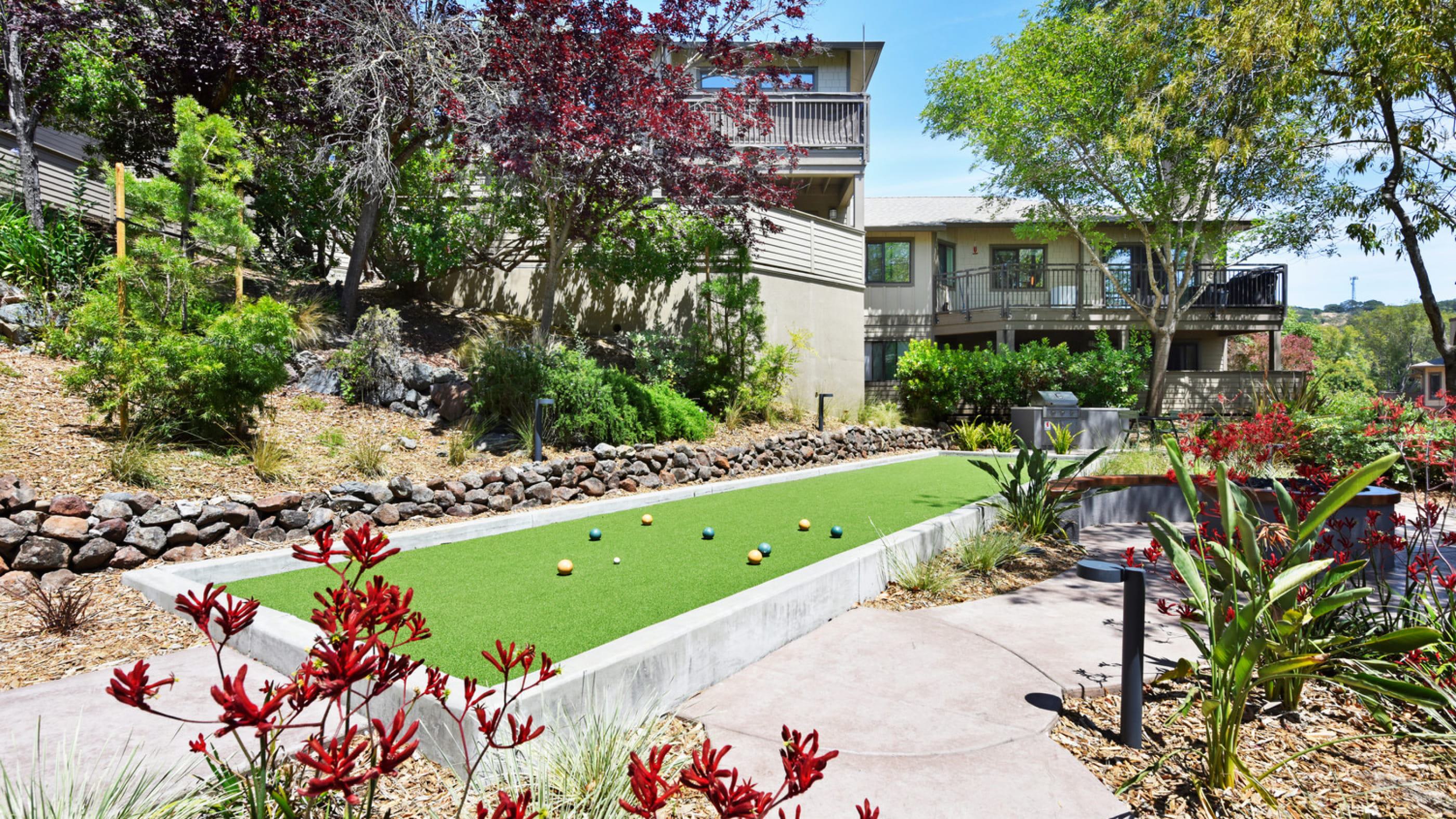 depicts a sunny, landscaped outdoor courtyard of a residential complex, featuring paved seating areas surrounded by natural greenery and a bocce ball court