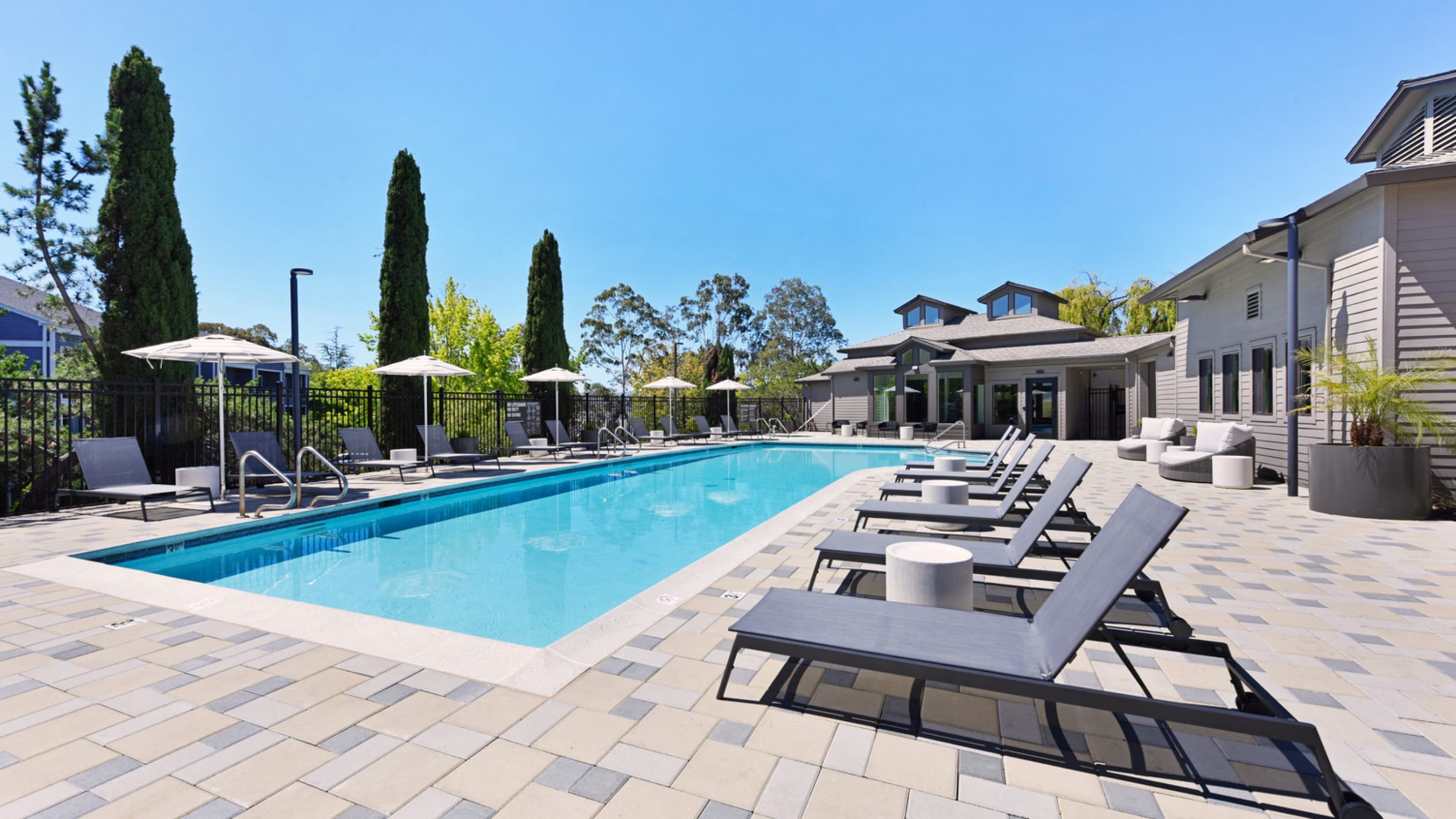 Displays a pristine outdoor swimming pool surrounded by a patterned paver deck, modern lounge seating, and patio umbrellas under a bright blue sky.