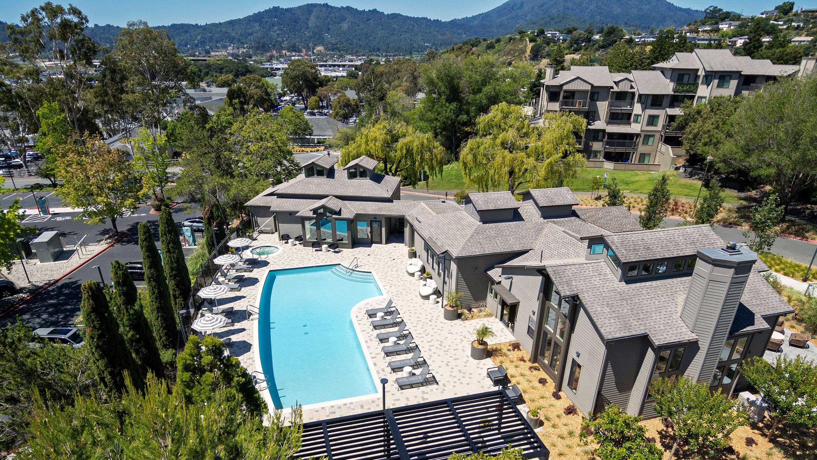 Displays a pristine outdoor swimming pool surrounded by a patterned paver deck, modern lounge seating, and patio umbrellas under a bright blue sky.