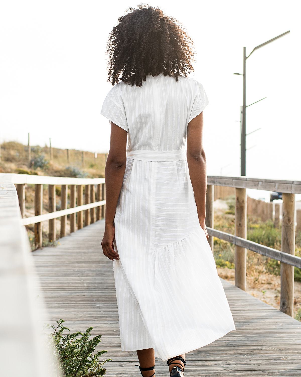 A woman with curly dark hair seen from behind, wearing a long white striped dress, walking along a wooden boardwalk in Larkspur, California. The sun is low, creating a warm glow, with coastal greenery and a wooden railing visible in the background.