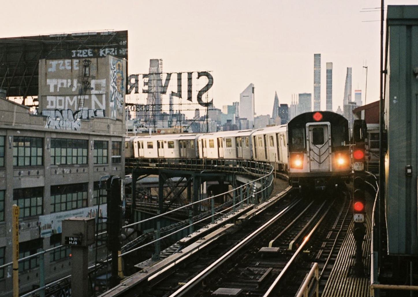 An elevated NYC subway train curves through Long Island City at sunset, passing industrial buildings and the iconic Silvercup Studios sign, with the Manhattan skyline rising in the distance.