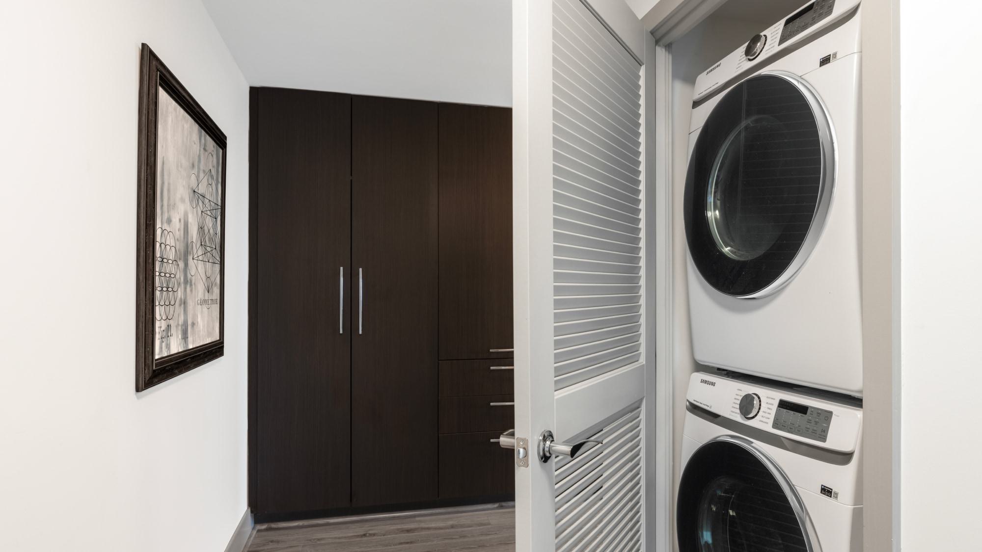 Full-size stacked Samsung washer and dryer inside apartment laundry closet with louvered door and modern wood cabinetry nearby.