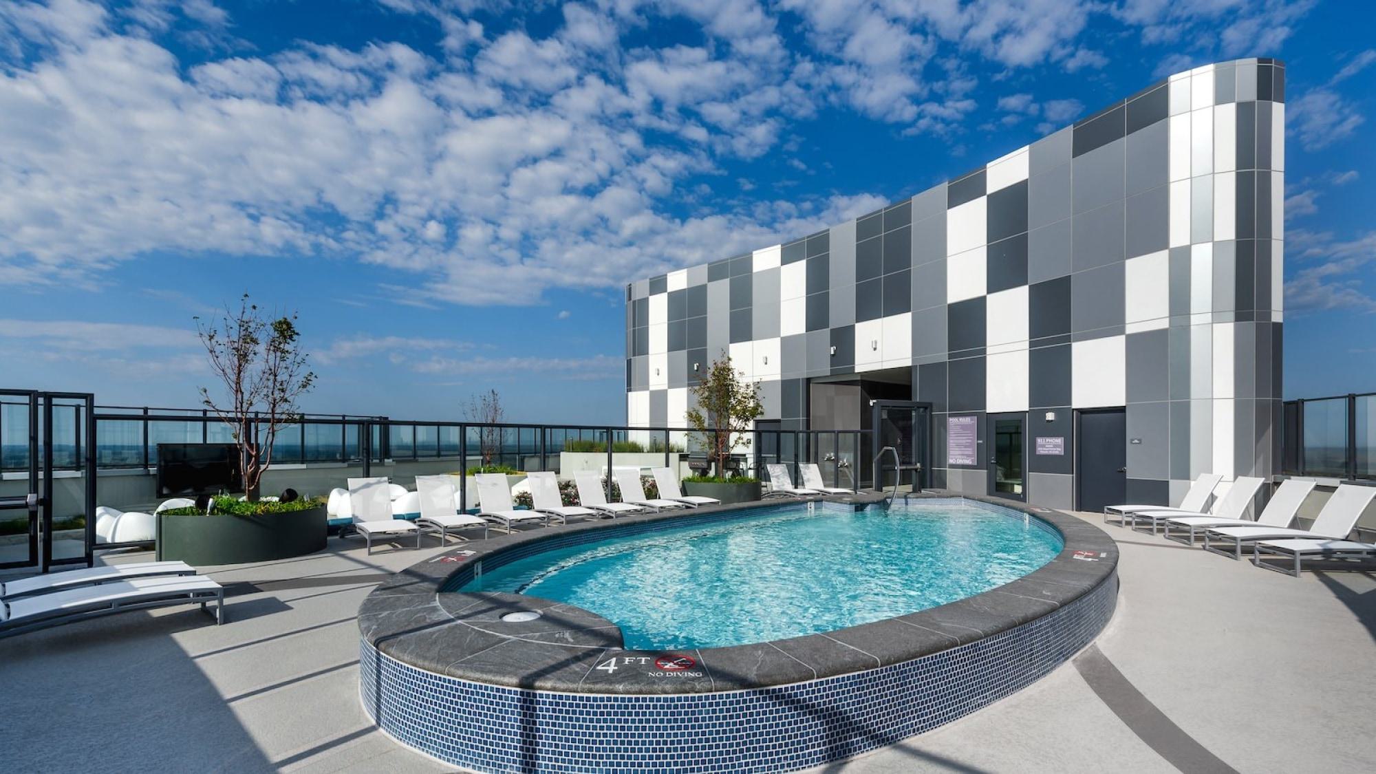 Curved rooftop pool at Adaire surrounded by white chaise lounges, glass guardrails, and open sky views, set against a modern checkerboard façade.