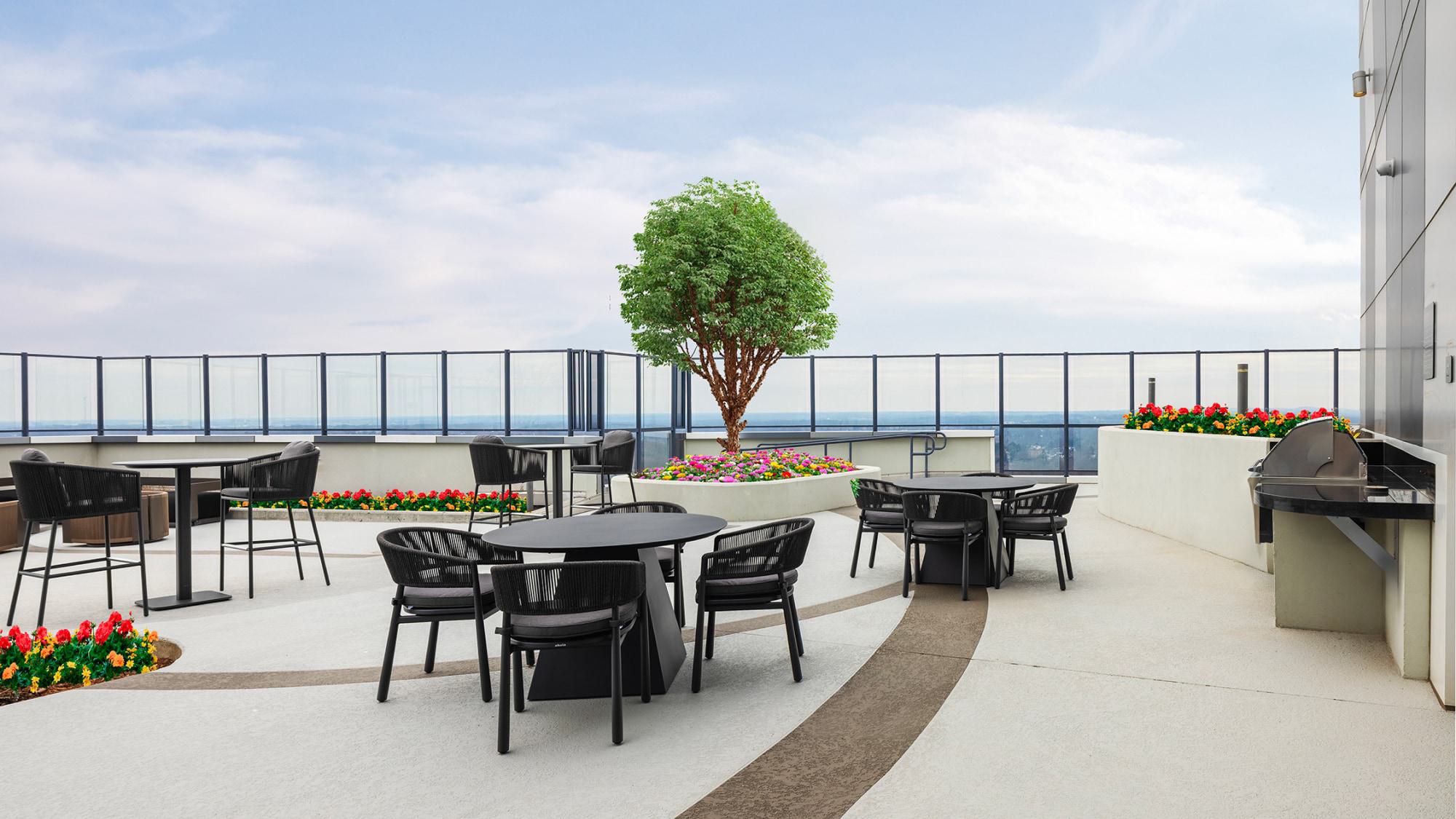 Rooftop terrace at Adaire featuring built-in grill station, black café tables with woven chairs, landscaped planters with flowering beds, and glass railings framing expansive skyline views.