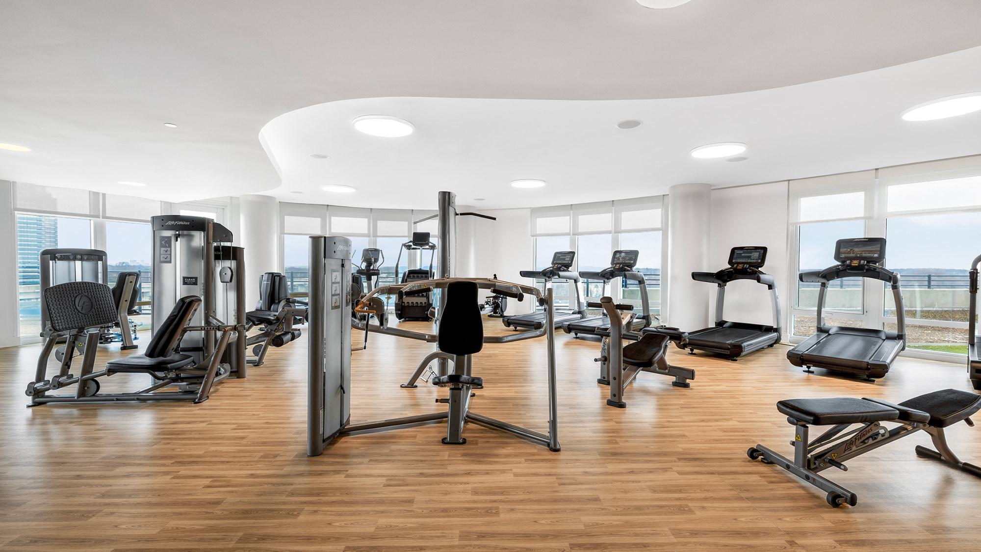 Bright, modern fitness center with wood floors, floor-to-ceiling windows, treadmills, strength machines, and free weights overlooking expansive views.