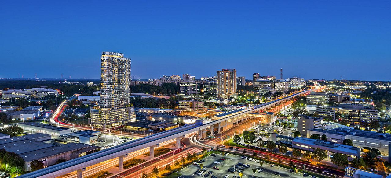 Aerial twilight view of Tysons, Virginia skyline with high-rise towers, illuminated offices, and the elevated Silver Line Metro tracks running above Route 7.