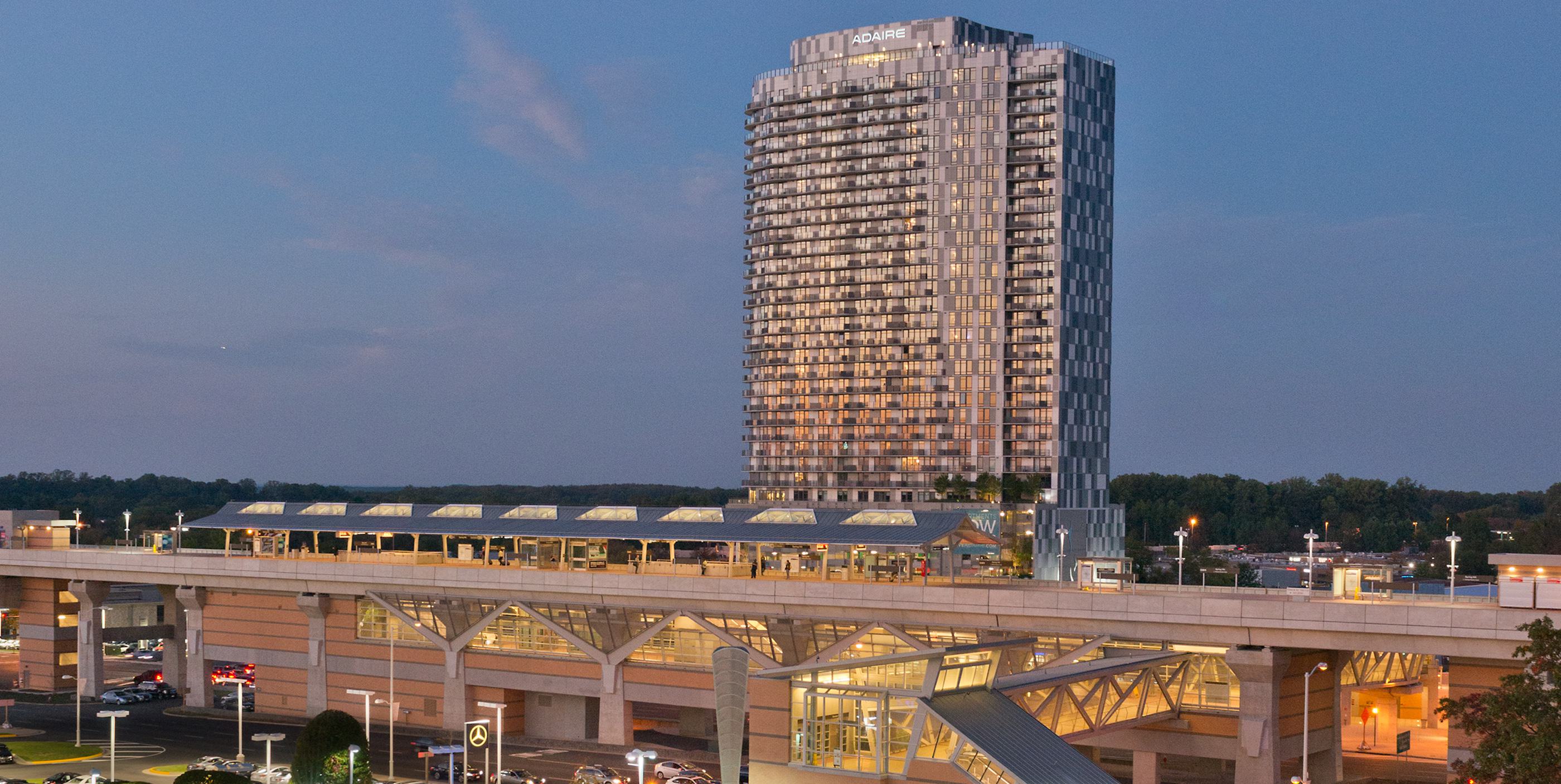 Elevated Spring Hill Metro station at dusk in Tysons, VA with Adaire’s high-rise tower illuminated in the background, platforms and pedestrian bridge visible below.