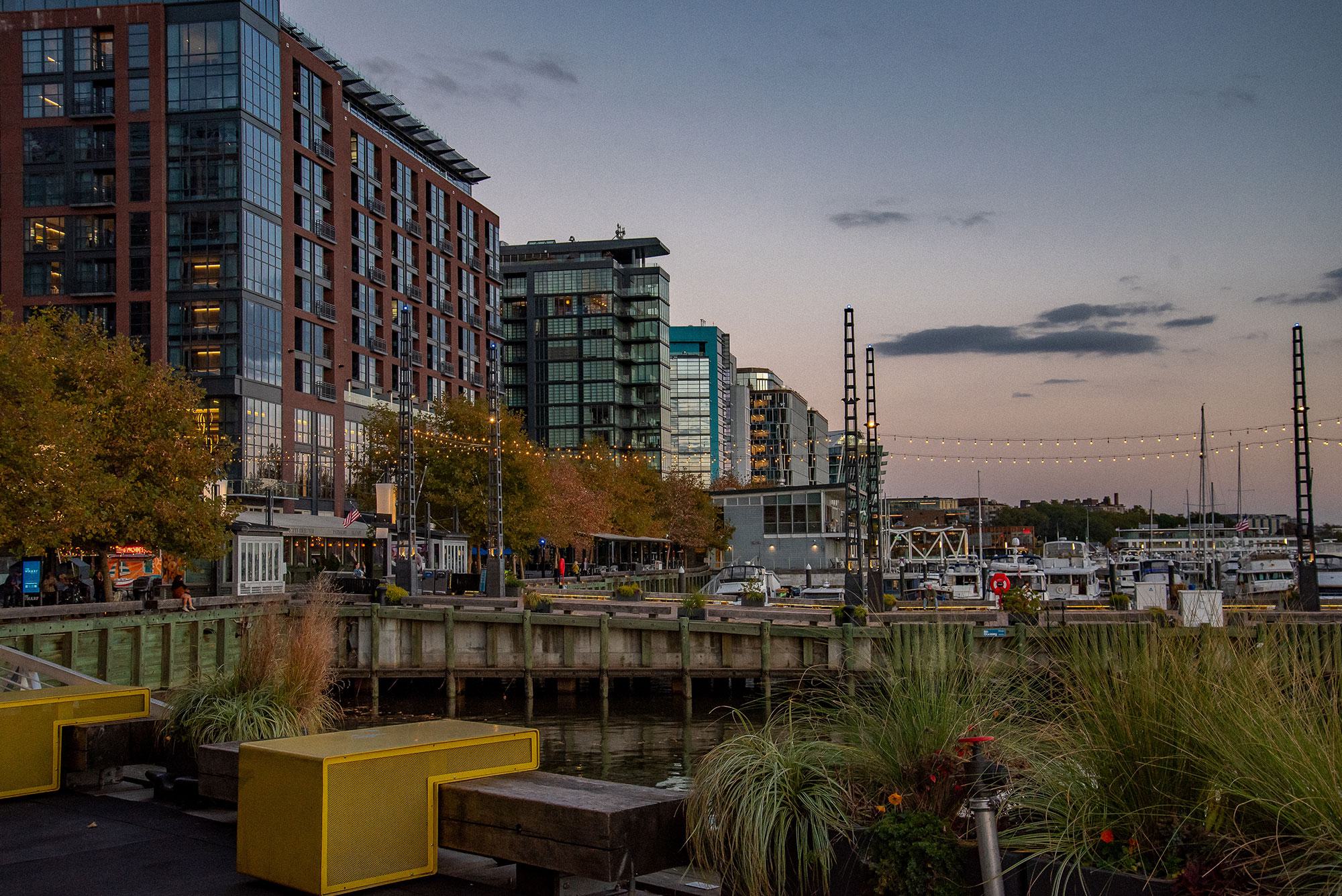 Evening view of The Wharf in Southwest DC, with waterfront apartments, docked boats, string lights, and riverwalk seating along the Washington Channel.