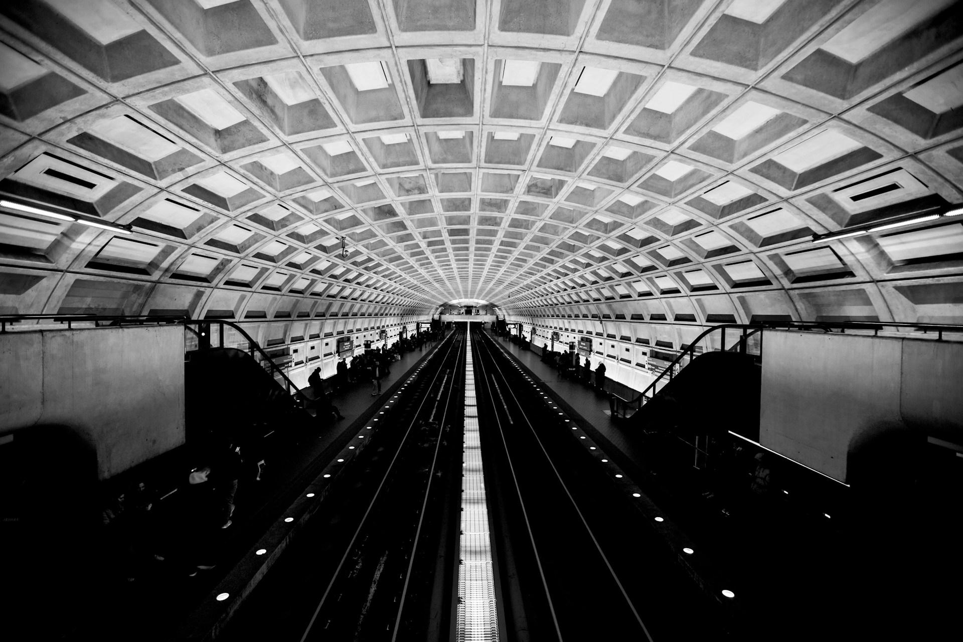Black-and-white photo of a Washington Metro station with a vaulted coffered concrete ceiling, twin tracks running down the center, and passengers waiting along the side platforms.