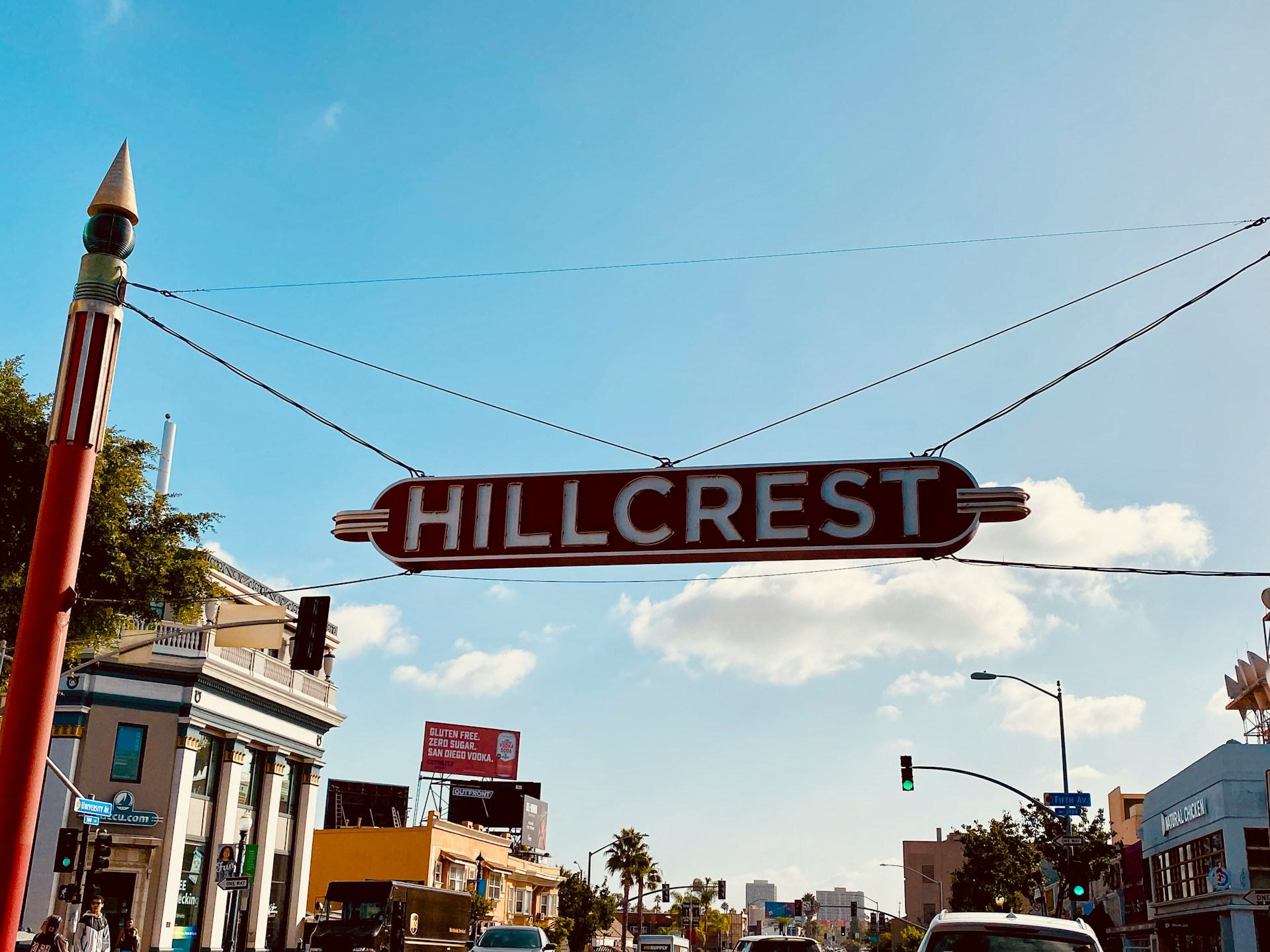 Hillcrest neighborhood sign suspended above University Avenue in San Diego, marking the entrance to the busy Uptown district with shops and traffic below.