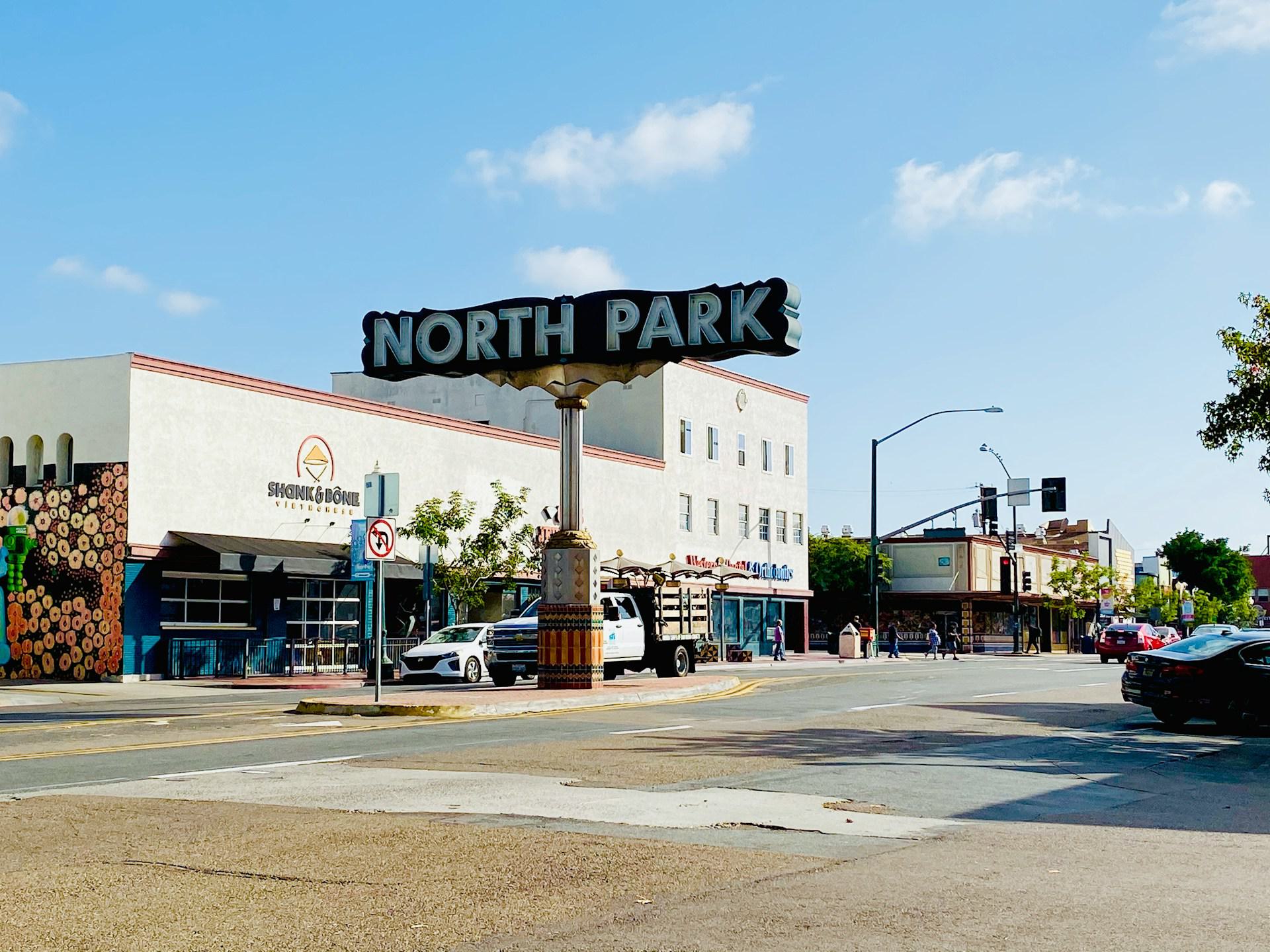 North Park neighborhood sign rising above the intersection of 30th Street and University Avenue in San Diego, with storefronts and clear blue sky.