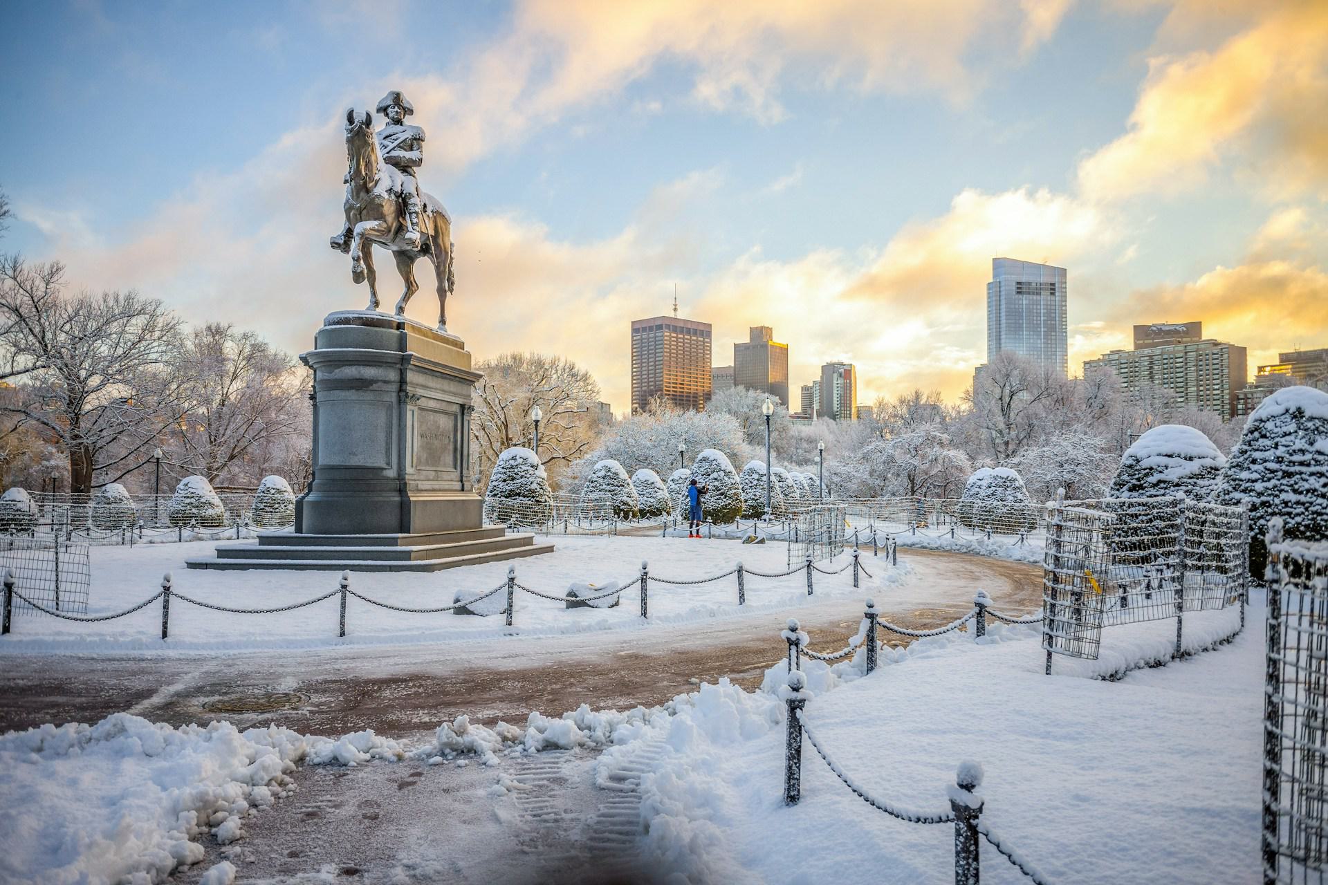 Snow-covered Boston Public Garden with the George Washington statue and downtown Boston skyline at sunrise.