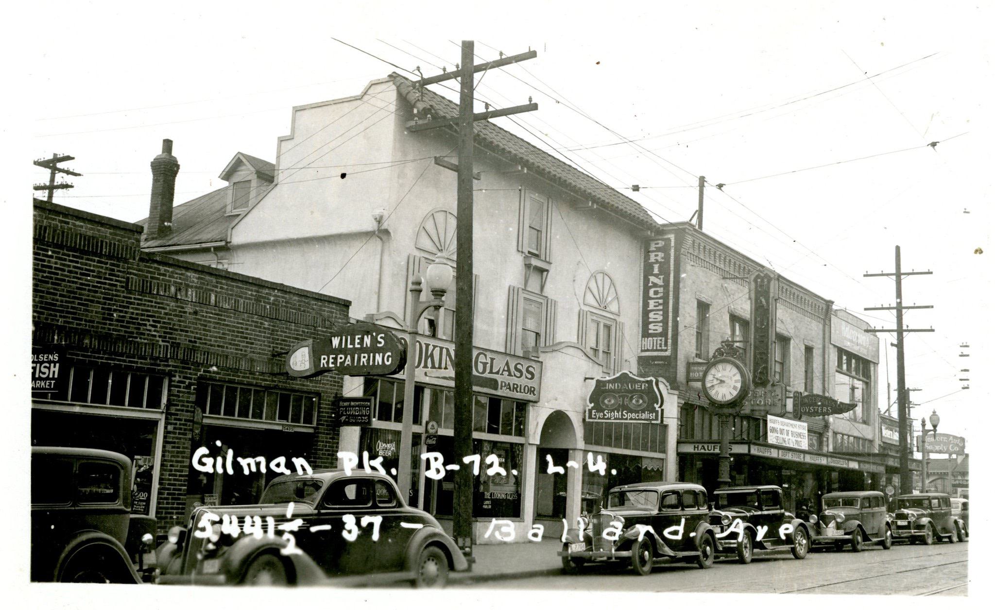 Historic black-and-white photo of Ballard Avenue with 1930s cars, storefronts, and streetcar lines in downtown Ballard.