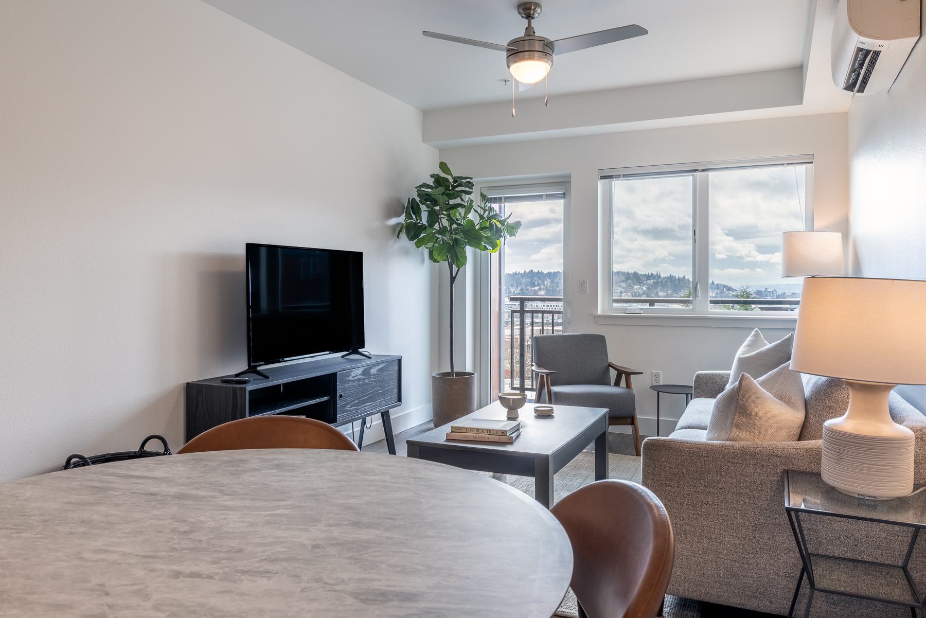 Living room at Urbana in Ballard with large windows, balcony door, air conditioning, and views across the Ballard neighborhood toward distant hills.