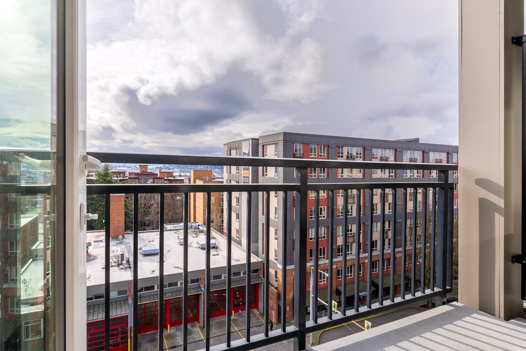 Private balcony at Urbana in Ballard overlooking nearby apartments, rooftops, and neighborhood streets with distant Puget Sound and hillside views.