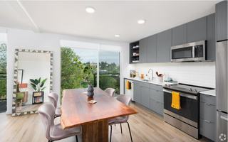 Bright modern kitchen with stainless steel appliances, quartz countertops, euro-style cabinetry, and large window opening to a balcony with tree-lined views.