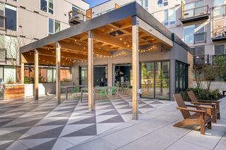 Courtyard pavilion at Arlo with wood pergola, string lights, outdoor dining tables, BBQ grill station, and indoor lounge connected by glass garage doors.