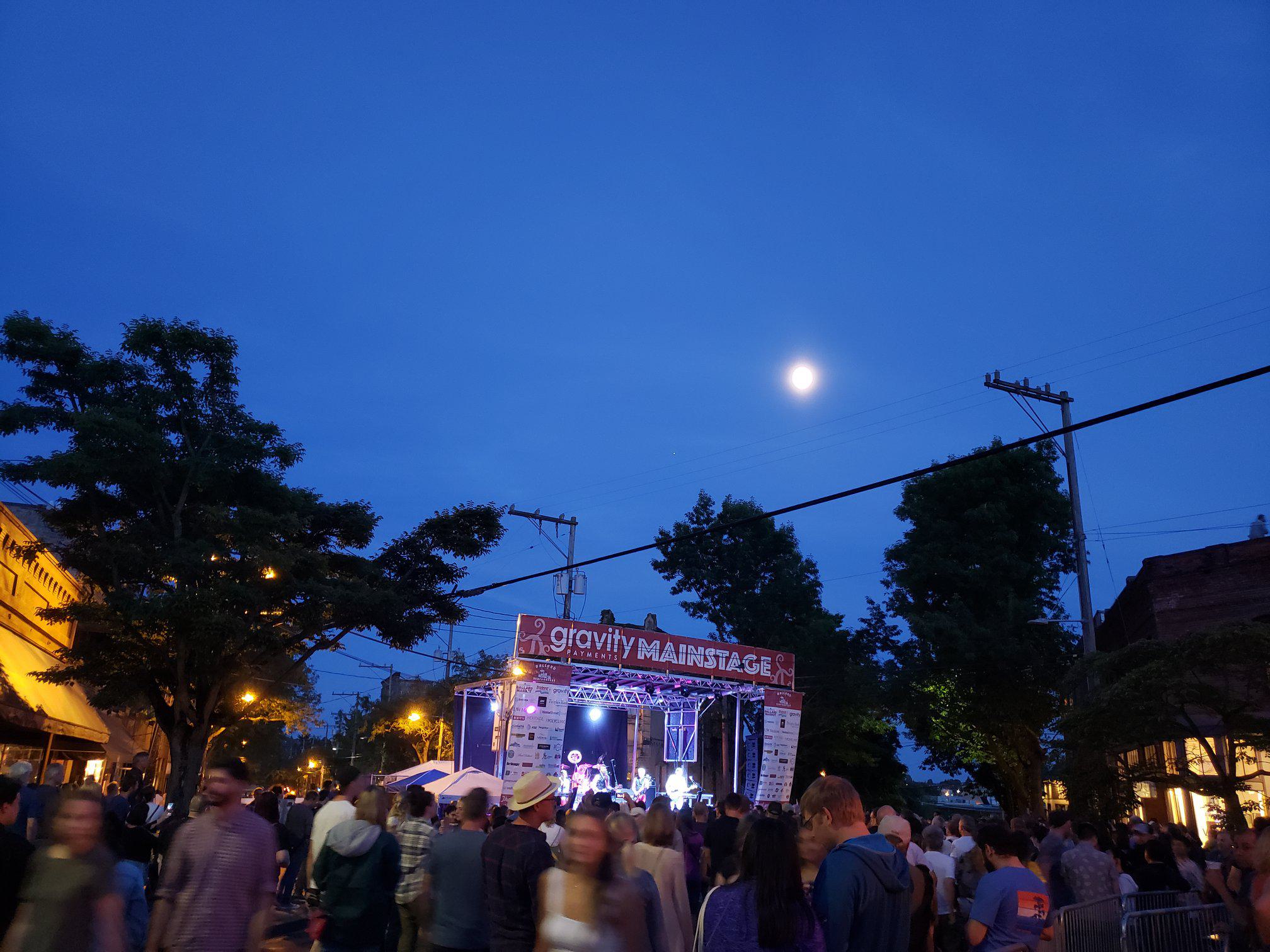Crowds gathered for a live music performance at the Ballard SeafoodFest main stage along NW Market Street at dusk, with festival lights glowing and the moon rising overhead.