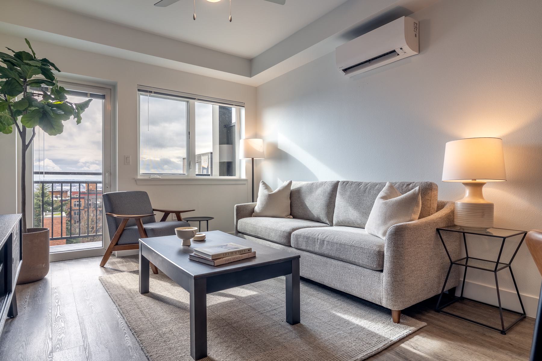 Living room at Urbana in Ballard with large windows, balcony access, ductless air conditioning unit, and views across nearby rooftops and neighborhood buildings.