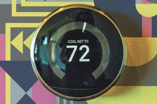 A circular Nest Learning Thermostat with a metallic finish mounted on a colorful geometric wall, displaying a digital screen set to cool at 72 degrees