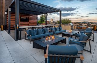 A wide-angle shot of The Hale’s rooftop terrace at dusk, featuring two modern linear fire pits surrounded by navy blue lounge sofas and armchairs. A large black pergola with an integrated TV and slatted wood wall sits in the background, alongside a stainless steel grilling station and outdoor dining tables, all overlooking the Washington D.C. skyline under a twilight sky.