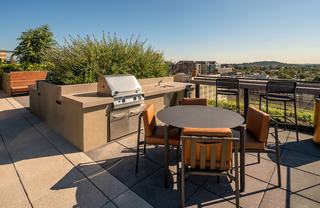 Modern outdoor rooftop lounge at The Hale in NoMa featuring a black steel pergola, wood-slat feature wall with a flat-screen TV, navy blue sofa seating, a large dining table, and professional stainless steel grilling stations under a clear blue sky.