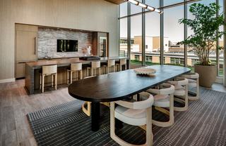 Modern indoor rooftop sky lounge at The Hale featuring a large oval dark wood dining table with curved cream chairs, a stone waterfall-edge island with bar seating, and a sleek catering kitchen with marble accents. Floor-to-ceiling windows offer views of the NoMa neighborhood under a warm, ambient light.