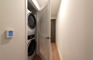 A laundry closet in a modern apartment at The Hale featuring a stacked set of full-size, front-loading washer and dryer units. The machines are tucked behind a white louvered door in a hallway with light wood-style flooring.