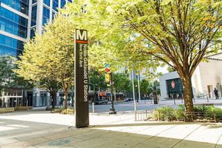 A tall, dark bronze Metro station pylon for the NoMa-Gallaudet U Station (New York Ave) stands on a sunlit sidewalk in Washington, D.C., framed by lush green and yellow autumn trees and modern glass office buildings.