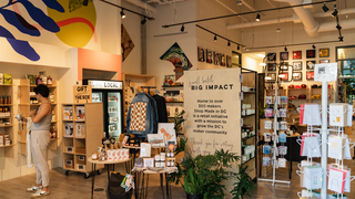 An interior view of the Shop Made in DC store in Union Market, featuring bright, modern shelving filled with locally made gifts, jewelry, and art. A large wooden sign in the center reads "Small Batch, Big Impact," highlighting the shop’s mission to support the D.C. maker community.