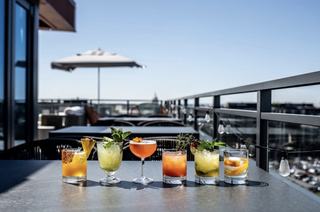 A stunning lineup of six colorful craft cocktails garnished with fresh fruit and herbs on a rooftop table at Sly. The sun-drenched terrace at The Morrow Hotel shows a blurred NoMa-Union Market skyline under a clear blue sky, capturing the upscale rooftop nightlife of Northeast D.C.