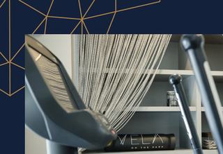 A close-up of a high-tech Expresso interactive bike at the Vela Cardio & Fitness Center. In the background, organized white shelving holds branded 'Vela on the Park' yoga mats and fitness equipment, framed by a modern geometric wall graphic