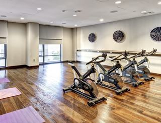 The modern Yoga and Wellness Studio at Vela featuring premium Stages spin bikes and a spacious wood-floored area for mat work. Large mirrors line the walls under soft lighting, with a flat-screen monitor for on-demand fitness classes and guided yoga sessions