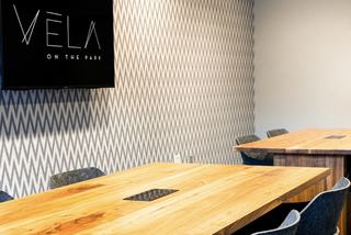 Modern coworking conference room at Vela on the Park featuring large live-edge wood tables with integrated power outlets, stylish blue chairs, and a chevron-patterned wall with a large display screen showing the property logo.