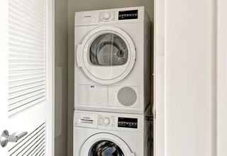 A bright, modern laundry closet at Vela on the Park featuring a stacked, white Bosch full-sized front-loading washer and dryer behind an open white louvered door.
