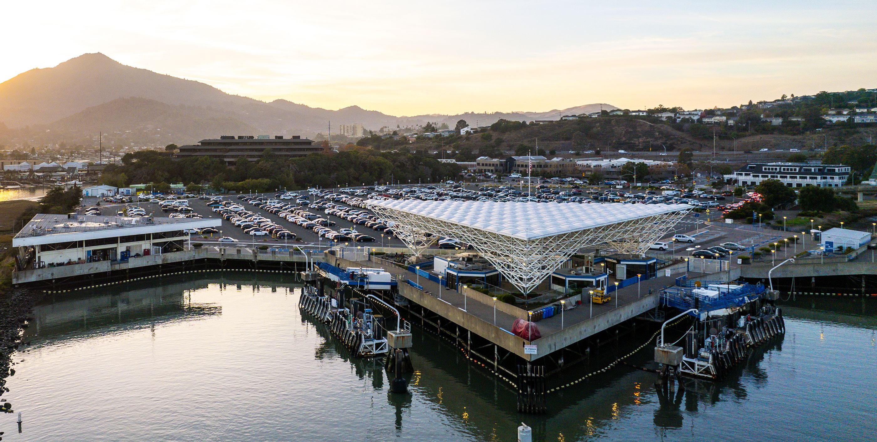 An aerial landscape view of the Larkspur Ferry Terminal at sunset. The iconic white space-frame roof of the terminal sits over the water, with a large parking lot and the marshy coastline nearby. In the background, the sun sets behind the silhouette of Mount Tamalpais under a clear sky with a single jet trail.