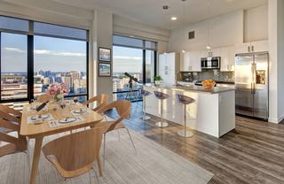 A wide-angle, eye-level shot captures a bright, modern open-concept penthouse featuring a dining area and kitchen with expansive city views.