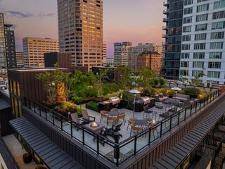 An elevated view of the rooftop terrace at The Victor during sunset, featuring a modern fire pit, outdoor grilling stations, dining tables, and lush landscaping against the Seattle skyline.