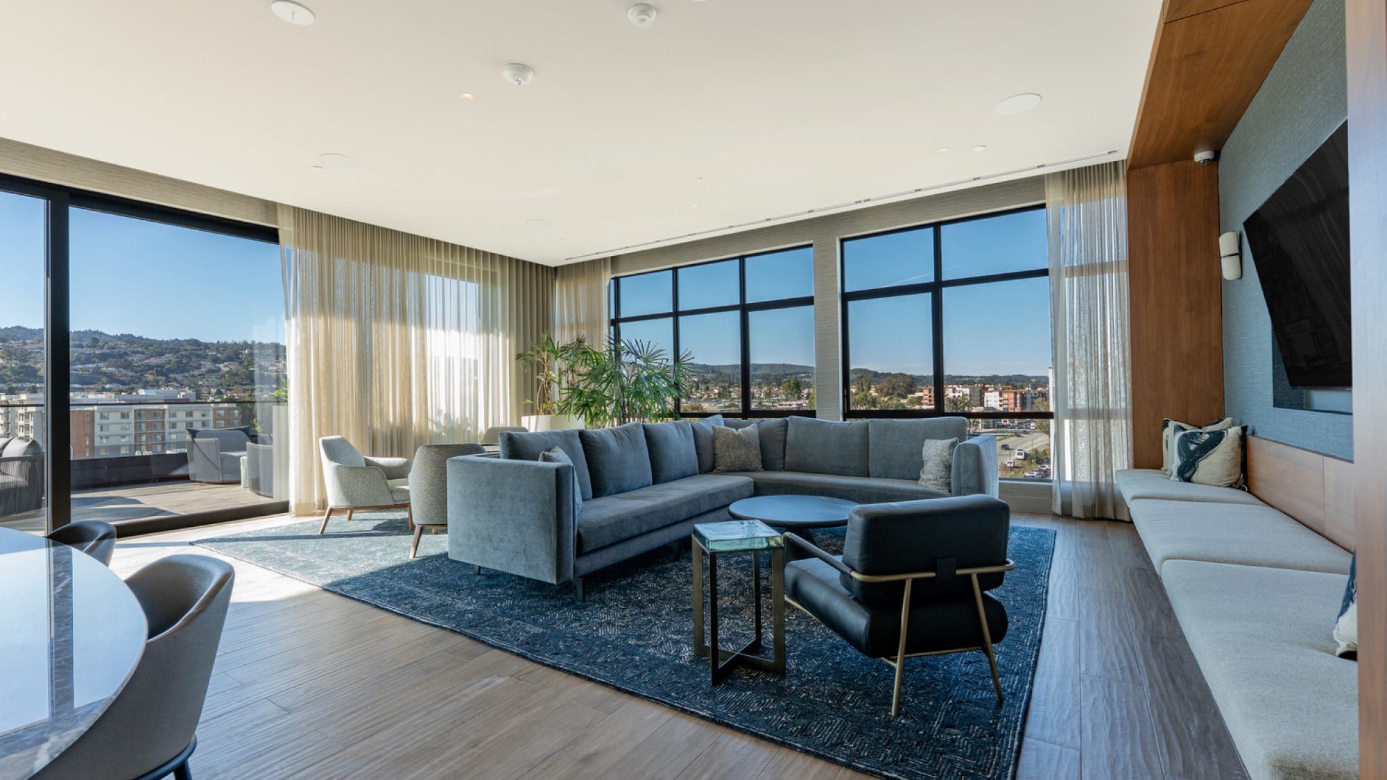 A high-ceilinged modern lounge with floor-to-ceiling corner windows overlooking the Burlingame cityscape and the Bay under a bright morning sky.