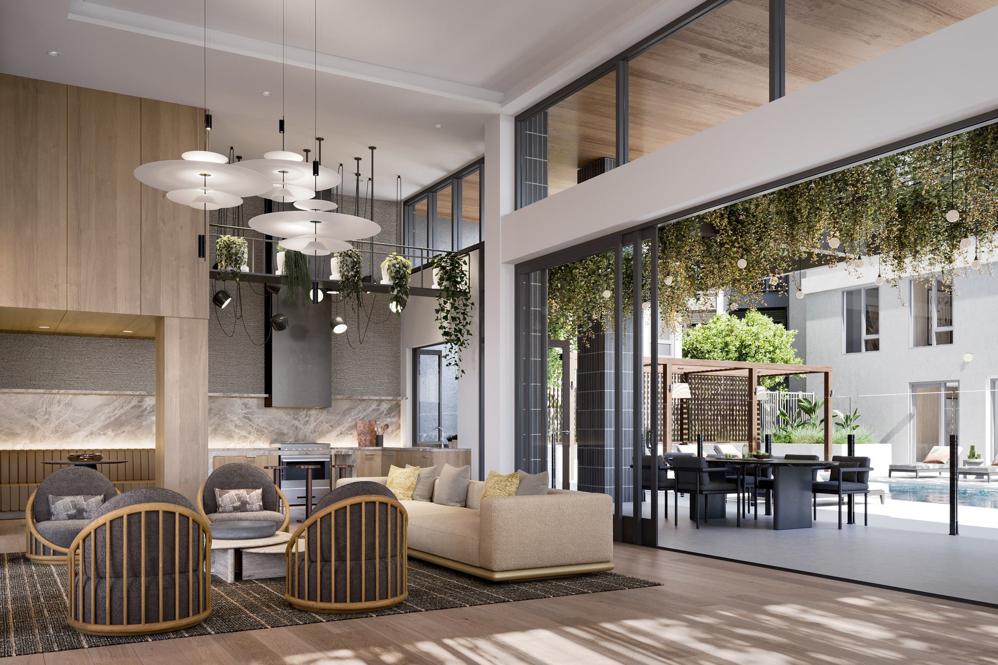 A high-ceilinged interior shot of the Revery demonstration kitchen and lounge flowing into an outdoor courtyard. The kitchen features marble backsplashes, light wood cabinetry, and a designer pendant light fixture. Massive black-framed glass doors are slid open, revealing an outdoor dining table under a pergola, lush hanging greenery, and a glimpse of the pool deck in the sunlit Zen Courtyard.