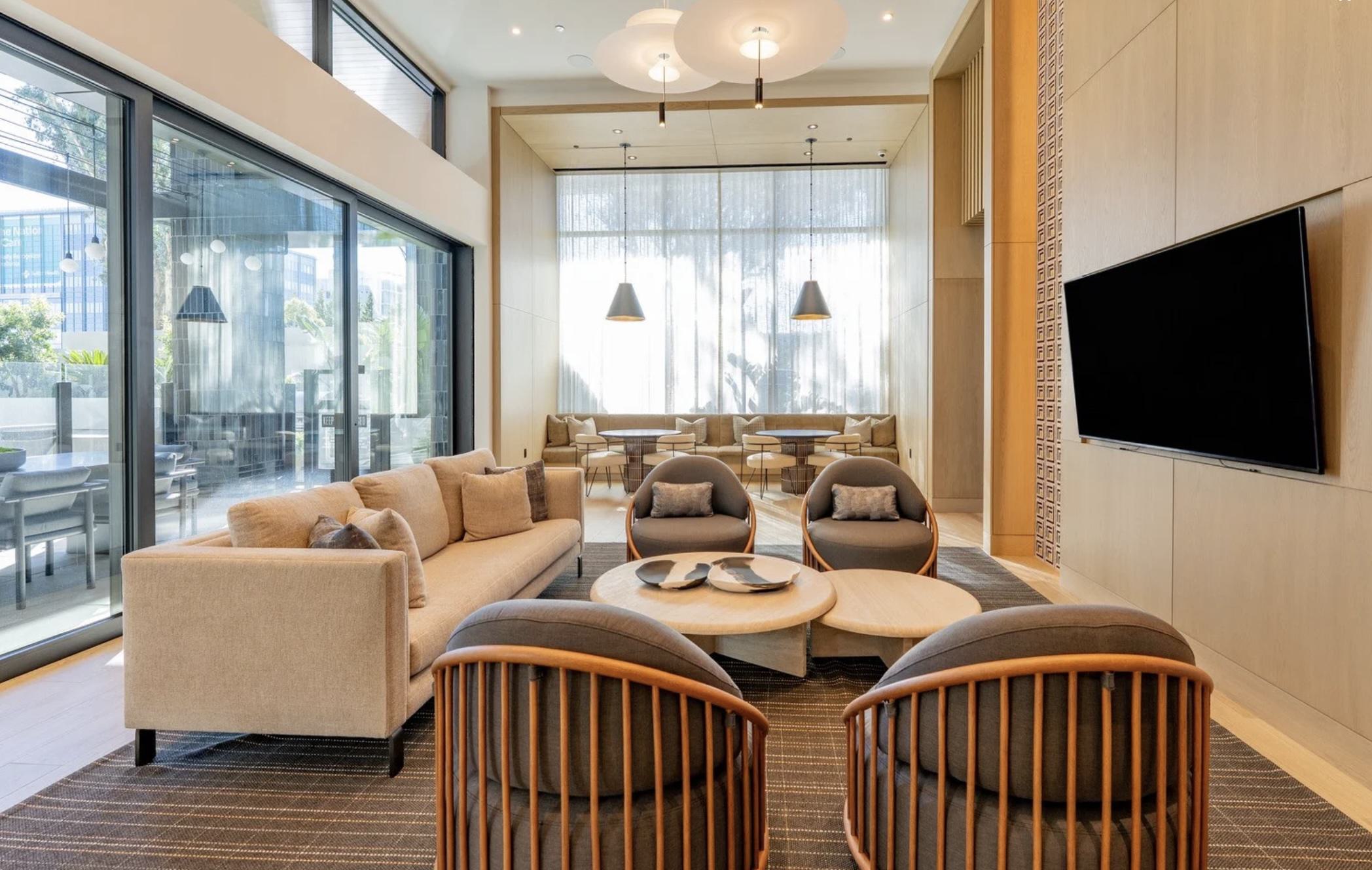 A bright, sun-drenched interior view of the Revery club lounge. The space features light wood wall paneling, a large wall-mounted television, and a neutral-toned sofa paired with modern, curved wooden-slat armchairs. In the background, a recessed dining nook with café tables and a long banquette is visible beneath large windows. To the left, an open doorway leads into a billiards room.
